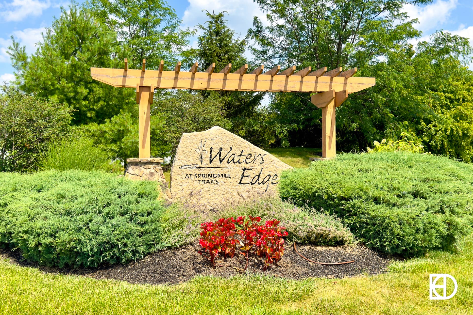 Neighborhood entrance sign for Waters Edge at Springmill Trails with stone etched with title, under wood pergola and landscaping.